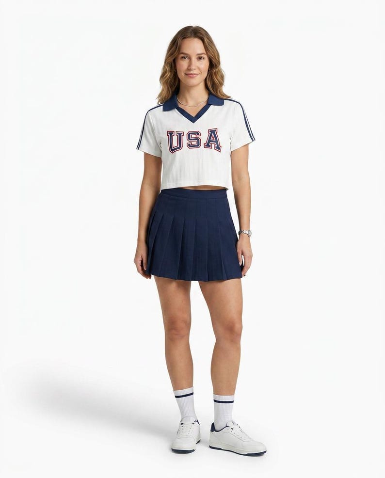 May include: A white short-sleeved cropped polo shirt with a navy blue collar and sleeve stripes, featuring the text USA in navy blue with red outlines. Paired with a navy blue pleated mini skirt, white crew socks with navy stripes, and white sneakers.