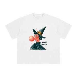 May include: A white t-shirt featuring a graphic illustration of a person wearing a black witch hat with a leopard print band. The person has dark hair, is blowing a large pink bubble, and is wearing a black cape. The text BASIC WITCH is printed in black.