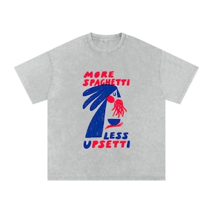 Può includere: Una t-shirt grigio chiaro a maniche corte con un'illustrazione grafica di una figura blu che mangia spaghetti rossi da una ciotola. Il testo recita MORE SPAGHETTI in rosso e LESS UPSETTI in blu. Il design ha un aspetto testurizzato.