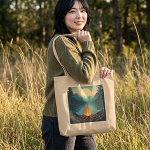 Könnte beinhalten: Beige Canvas-Tragetasche mit einer Naturszene mit Lagerfeuer, Bergen und Sonnenuntergang. Die Tasche hat lange Träger und wird von einer Person in einem grünen Pullover gehalten, die in hohem Gras steht.