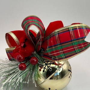 May include: A large gold-colored metal jingle bell ornament featuring snowflake-shaped cutouts. It is topped with a decorative bow made of red velvet and red, green, and gold plaid ribbon, accented with frosted pine needles and shiny red berries.