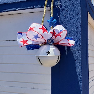 May include: A white metal jingle bell ornament featuring a decorative bow with red and blue star patterns. The ornament is suspended by a gold rope and includes a small, glittery blue star accent attached to the top of the bow for decoration.