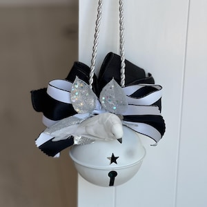 May include: A decorative white bell with a star cutout and keyhole design, adorned with a black and white striped bow, silver glitter accents, and a small white bird. The bell hangs from a silver rope.