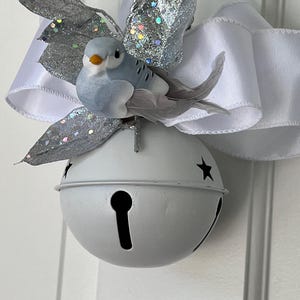 May include: A white metal jingle bell ornament featuring a light blue bird with grey feather wings perched on top. The ornament is decorated with a large white satin ribbon bow and silver glittered leaves, all suspended by a silver twisted cord.