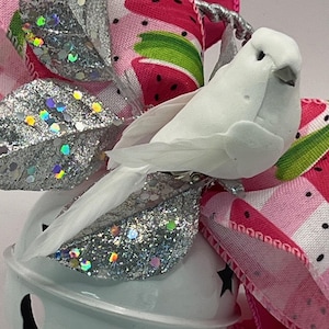 May include: A white bird figurine with feathered wings perched atop a white jingle bell. The bird is accented by silver glittered leaves and a decorative ribbon featuring a pink and green watermelon slice pattern with black seeds.