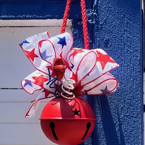 May include: A large red metal jingle bell ornament featuring a star-shaped cutout and vertical slits. It is topped with a decorative bow made of white ribbon with red and blue glitter stars, accented by red, white, and blue wire spirals and a red rope hanger.