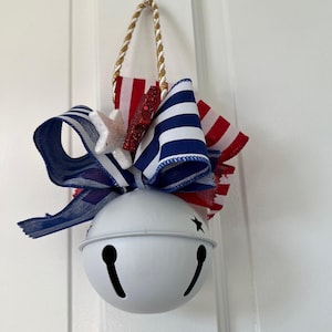 May include: A white metal bell with a patriotic theme, featuring a red, white, and blue ribbon and star accents. The bell has a gold rope for hanging and is decorated with a bow made of striped and glittery fabric. A festive decoration for the home.