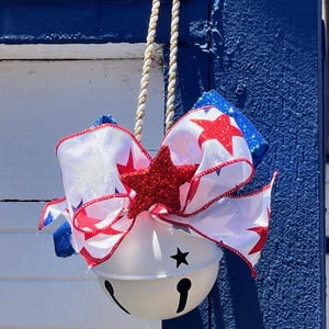 May include: A white jingle bell ornament hanging from a twisted gold and white cord. The bell is topped with a large decorative bow featuring white fabric with red stars, blue glitter accents, and a prominent red glitter star in the center.
