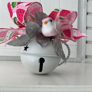 May include: A white bell ornament with a keyhole design, adorned with a pink and white watermelon-patterned bow and silver glitter leaves. A small white bird with a pink head sits atop the bell. A star cutout is visible on the bell.