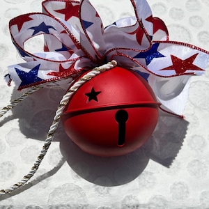 May include: A matte red spherical jingle bell featuring a star-shaped cutout and a vertical slit. The bell is adorned with a white ribbon patterned with red and blue stars, tied into a bow, and includes a twisted white and gold cord for hanging.