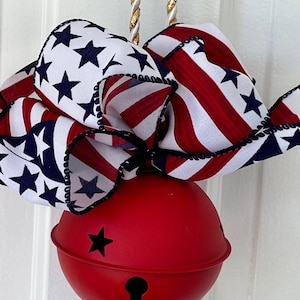 May include: A red bell with a star-shaped cutout and a keyhole design, topped with a patriotic ribbon featuring red and white stripes and blue stars. The bell hangs from a gold and white rope.
