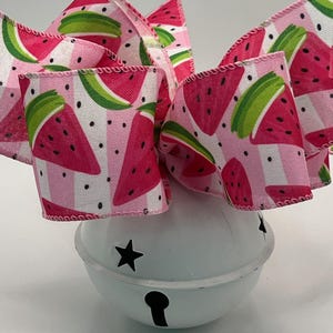 May include: A white metal jingle bell with star-shaped cutouts and a central vertical slit, topped with a large, multi-looped fabric bow featuring a repeating pattern of pink watermelon slices, green rinds, and black seeds on a striped background.