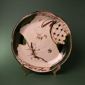 May include: A round ceramic plate with a cream-colored base featuring hand-painted dark brown botanical motifs and a grid pattern. The rim is decorated with a wavy brown line and accented with deep green glaze patches along the scalloped edge.