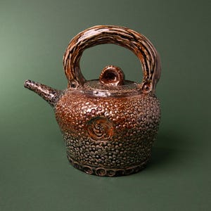 Może przedstawiać: Ceramiczny czajniczek o teksturowanej, ziarnistej powierzchni w odcieniach brązu i szarości. Posiada wyraźny, gruby, łukowaty uchwyt o wyglądzie drewna, mały dzióbek oraz pokrywkę z centralnie umieszczonym uchwytem w kształcie gałki.