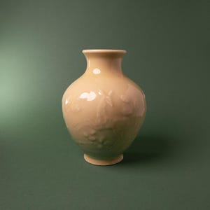 May include: A pale peach-colored ceramic vase with a rounded body and a flared rim. The surface features a subtle, raised relief design depicting a deer standing among foliage, integrated into the smooth, glossy finish of the vessel.
