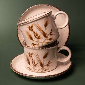 May include: Two off-white ceramic cups and two matching saucers stacked vertically. The pieces feature a speckled finish, brown rim detailing, and a brown botanical illustration of wheat stalks on the exterior of each cup.