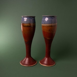 May include: Two tall ceramic goblets featuring a multi-toned glaze. The upper section is a soft blue, transitioning into bands of tan, brown, and deep red toward the base. Each vessel has a flared rim and a slender stem leading to a wide foot.