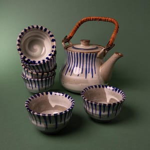 Puede incluir: Un juego de té de cerámica que incluye una tetera con asa de ratán curvada y cinco tazas a juego. Las piezas son de color blanco roto con textura moteada y están decoradas con patrones de pinceladas azules verticales en los bordes.