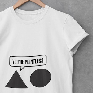 Puede incluir: Camiseta blanca con un diseño gráfico. El diseño presenta un bocadillo con el texto "YOU'RE POINTLESS" encima de un triángulo negro y un círculo negro. La camiseta está colgada de una percha de madera.