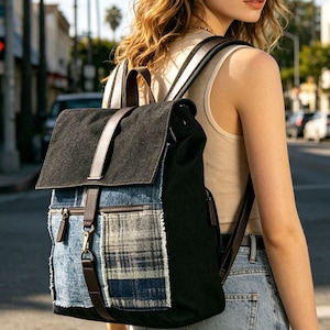 May include: A backpack featuring a black top flap and side panels, with a front section composed of patchwork denim and plaid fabric panels. The bag includes a central brown leather-like strap with a metal clasp and adjustable shoulder straps.