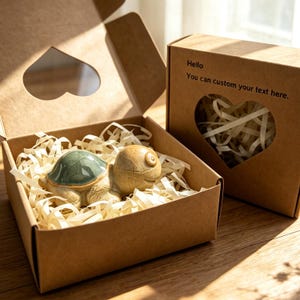 May include: A small ceramic turtle figurine with a green shell and tan body nestled in a brown cardboard gift box filled with shredded paper. The box lid features a heart-shaped cutout and the text Hello You can custom your text here.