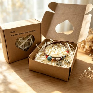 Puede incluir: Una caja de regalo de cartón marrón con un recorte en forma de corazón que contiene una pulsera de cuentas con un dije de mariposa y hoja de bronce. La tapa tiene el texto Hello You can custom your text here. La pulsera descansa sobre papel.