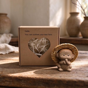 May include: A brown cardboard gift box with a heart-shaped cutout filled with white paper shreds, featuring the printed text 'Hallo' and 'You can custom your text here.' Beside the box sits a small, speckled ceramic monkey figurine wearing a straw hat.
