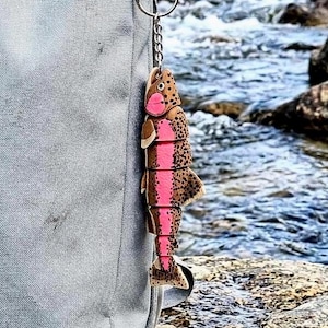 May include: A segmented trout keychain featuring a brown body with black spots and a prominent pink stripe along the side. The fish figure is attached to a silver metal keyring and chain, hanging vertically against a light gray fabric surface.