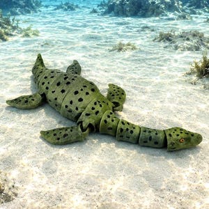 May include: A green and black spotted sea creature toy rests on a sandy ocean floor. The toy has a segmented body, flipper-like appendages, and a small head. The background shows rocks and clear blue water.