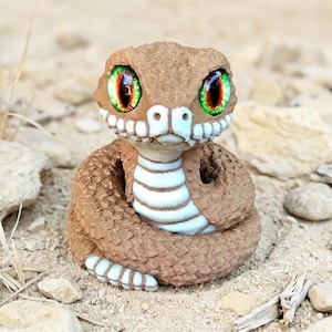 May include: A detailed, brown figurine of a coiled snake with large, expressive eyes. The snake has a textured body, white and blue accents, and is set against a sandy background with rocks and dry grass. The eyes are green with red highlights.