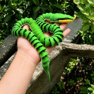 May include: A bright green toy snake with a yellow and black head is held in a hand. The snake is segmented, with a textured, scale-like appearance. The background features lush green foliage.