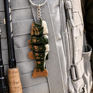May include: A segmented fish keychain featuring a dark green body with tan mottled spots, a light tan tail, and white underbelly sections. The fish is attached to a silver metal split ring and chain, hanging against a grey tactical backpack.