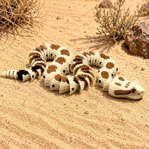 May include: A crocheted snake toy in a desert setting. The snake is cream-colored with brown spots and segments. The head is detailed with brown and black accents. The toy is lying on sand, with desert plants and rocks in the background.