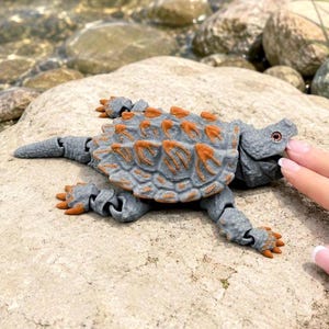 May include: A gray and orange toy dinosaur, possibly an Ankylosaurus, is resting on a rock near water. The toy has a textured, armored back with orange spikes and articulated limbs. The dinosaur's tail is extended, and a finger is touching its head.