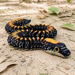 May include: A detailed, handcrafted sculpture of a snake with a segmented body. The snake features a black base color with orange and white accents. The sculpture is lying on a sandy surface with leaves.