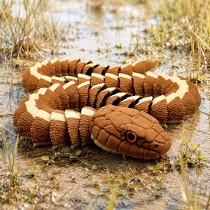 May include: A brown and cream-colored articulated snake toy resting on a muddy surface with grass. The snake's segmented body and head are detailed, with a textured pattern. The snake's eye is a dark brown color.