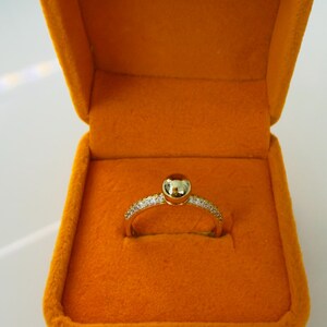 May include: A gold ring with a round, light-colored stone set in the center, surrounded by small, sparkling stones. The ring is displayed in an open, orange velvet ring box. The box is a bright, solid orange color.