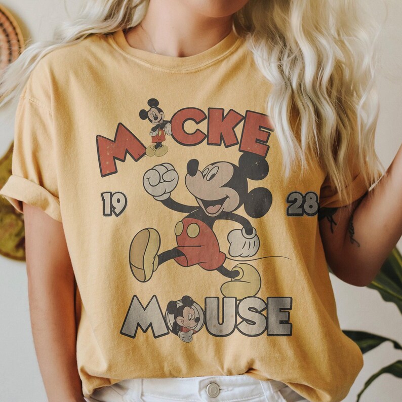 Op de afbeelding: Mosterdgeel T-shirt met een vintage Mickey Mouse-afbeelding. Het ontwerp bevat de woorden "Mickey Mouse" en de cijfers "19" en "28". Mickey Mouse wordt afgebeeld in een retro cartoonstijl.