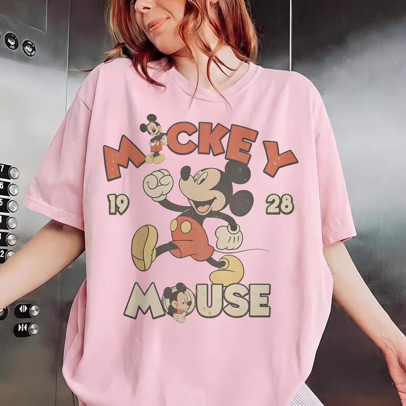 Op de afbeelding: Een lichtroze t-shirt met een vintage Mickey Mouse-ontwerp. Het ontwerp bevat de woorden "Mickey Mouse" en de cijfers "19" en "28". Mickey Mouse wordt afgebeeld in een rennende pose, met een rode broek en gele schoenen.