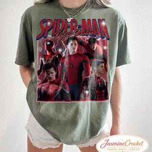以下が含まれることがあります： オリーブグリーンのTシャツ。赤と青で「Spider-Man Peter Parker」の文字とスパイダーマンのグラフィックがプリントされています。グラフィックにはスパイダーマンの複数の画像が含まれています。白いショートパンツと合わせています。