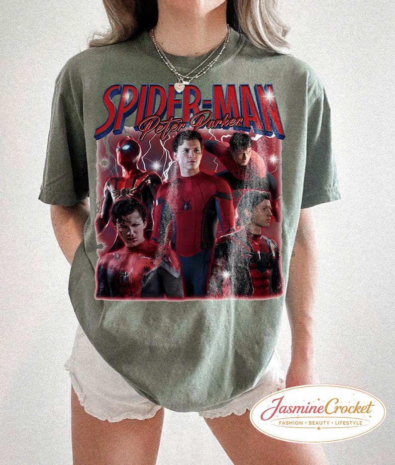 以下が含まれることがあります： オリーブグリーンのTシャツ。赤と青で「Spider-Man Peter Parker」の文字とスパイダーマンのグラフィックがプリントされています。グラフィックにはスパイダーマンの複数の画像が含まれています。白いショートパンツと合わせています。