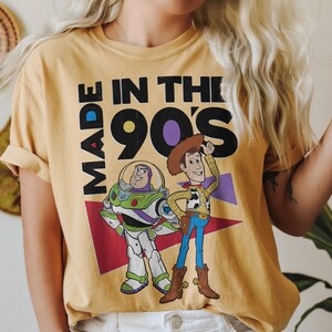 May include: Mustard yellow t-shirt featuring the text "MADE IN THE 90'S" and cartoon characters Buzz Lightyear and Woody from Toy Story. Buzz is in a green and white spacesuit, and Woody wears a cowboy hat and boots.