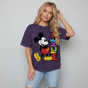 May include: A purple t-shirt with a Mickey Mouse graphic and the word "MICKEY" in colourful letters. The cartoon character is in his classic pose. The t-shirt has short sleeves and a crew neck. Light blue jeans are also visible.
