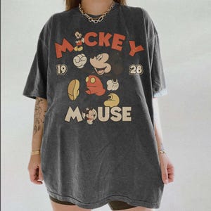 Op de afbeelding: Donkergrijze oversized t-shirt met een vintage Mickey Mouse-afbeelding. Het ontwerp bevat het woord "Mickey" in rood en "Mouse" in cr&egrave;me, samen met cartoonafbeeldingen van Mickey Mouse. Het shirt heeft een klassieke, versleten uitstraling.