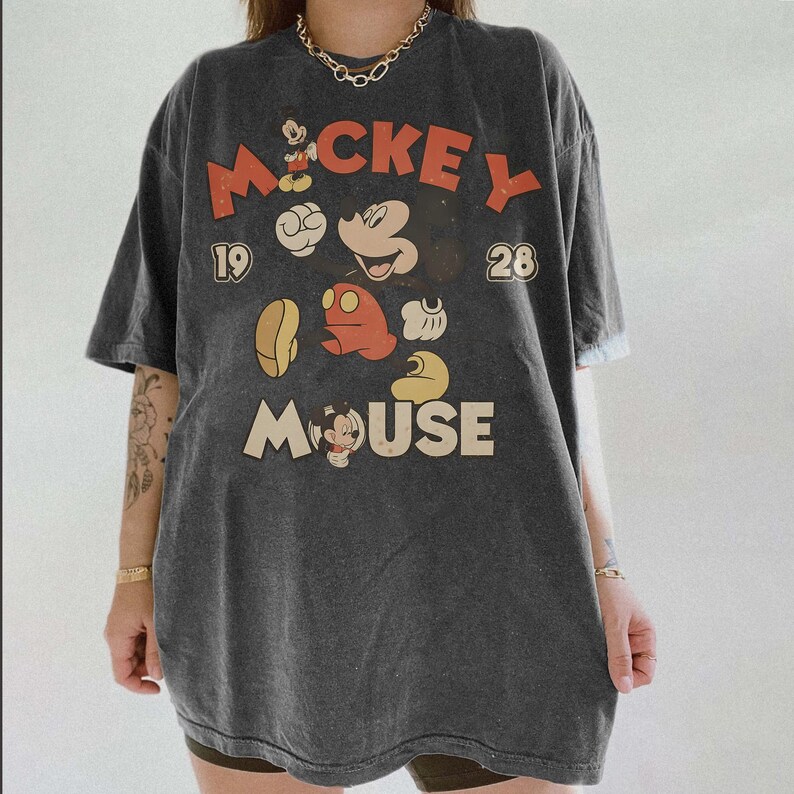 Op de afbeelding: Donkergrijze oversized t-shirt met een vintage Mickey Mouse-afbeelding. Het ontwerp bevat het woord "Mickey" in rood en "Mouse" in cr&egrave;me, samen met cartoonafbeeldingen van Mickey Mouse. Het shirt heeft een klassieke, versleten uitstraling.