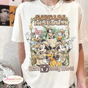 Op de afbeelding: Cr&egrave;mekleurig T-shirt met een afbeelding van Disney-personages in safari-outfit, waaronder Mickey Mouse, Donald Duck en Pluto. De tekst "ANIMAL KINGDOM" en "Walt Disney World" staan boven en onder de personages.