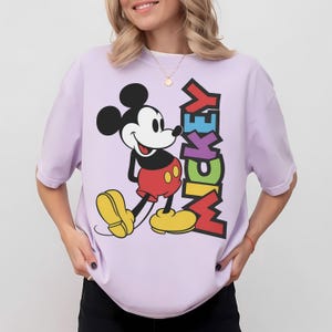 May include: A lilac t-shirt with a graphic of Mickey Mouse. The word "MICKEY" is printed vertically in colourful letters. Mickey is in his classic pose, wearing red shorts, yellow shoes, and black gloves.