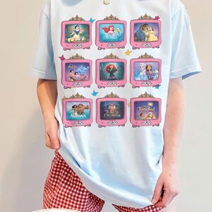 Op de afbeelding: Lichtblauw T-shirt met negen roze televisieschermen, elk met een andere animatiefilmsc&egrave;ne. Het shirt bevat sc&egrave;nes uit Sneeuwwitje, De Kleine Zeemeermin, Belle en het Beest en meer. De merknaam "Jasmine Crocket" staat onderaan.