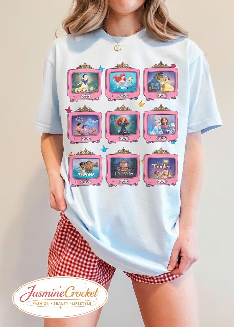 Op de afbeelding: Lichtblauw T-shirt met negen roze televisieschermen, elk met een andere animatiefilmsc&egrave;ne. Het shirt bevat sc&egrave;nes uit Sneeuwwitje, De Kleine Zeemeermin, Belle en het Beest en meer. De merknaam "Jasmine Crocket" staat onderaan.