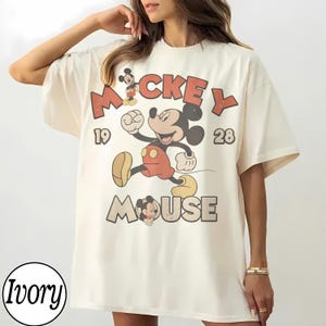 Op de afbeelding: Ivoor kleurig T-shirt met een vintage Mickey Mouse afbeelding. Het ontwerp bevat de woorden "Mickey Mouse" en de cijfers "1928". Mickey wordt afgebeeld in een rennende pose, met een rode broek en gele schoenen.
